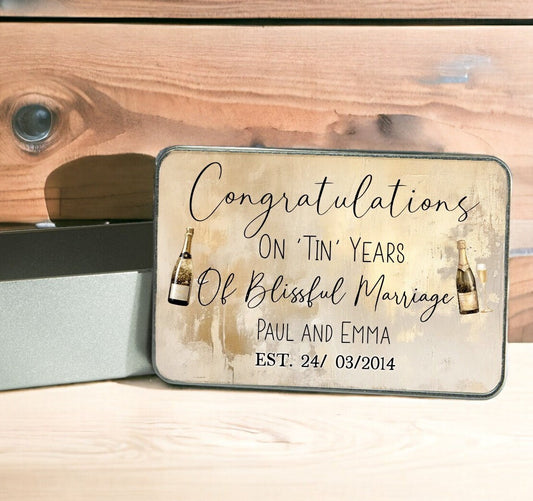 Rectangular silver tin with its lid off. The design on the tin lid has a gold background and the text reads Congratulations on Tin Years of Marriage, then the couples name and date. There are 2 champagne bottles on the design.