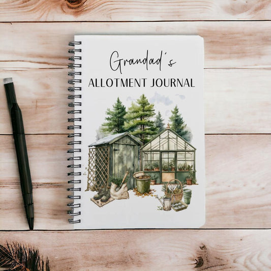 An A5 Lined Notebook that is spiral bound with an acrylic front and back cover which the design is printed onto. The design is a watercolour allotment space with a greenhouse, shed, trees and plants. the text reads Grandads Allotment Journal.
